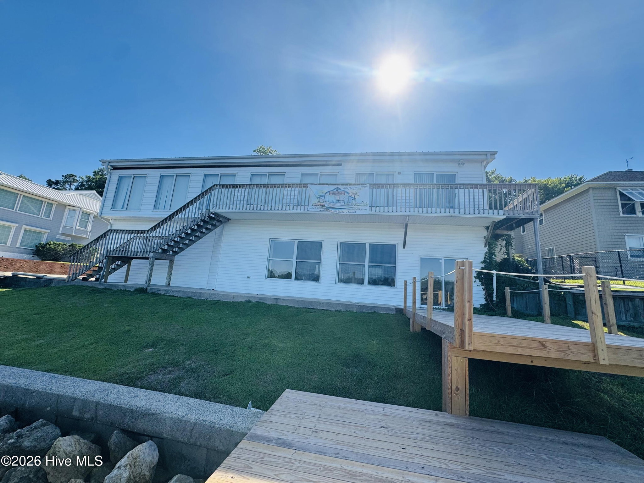 Water side of 888 Sea Gate Drive, a white two-story rear elevation with a full-width upper deck and exterior staircase, the back lawn running down to a rock seawall and wooden dock on the waterway