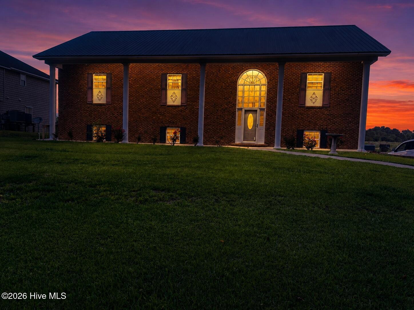888 Sea Gate Drive at twilight, a two-story brick manor with a full-height white-columned portico and arched fanlight, its windows glowing gold beneath a purple and orange sunset sky